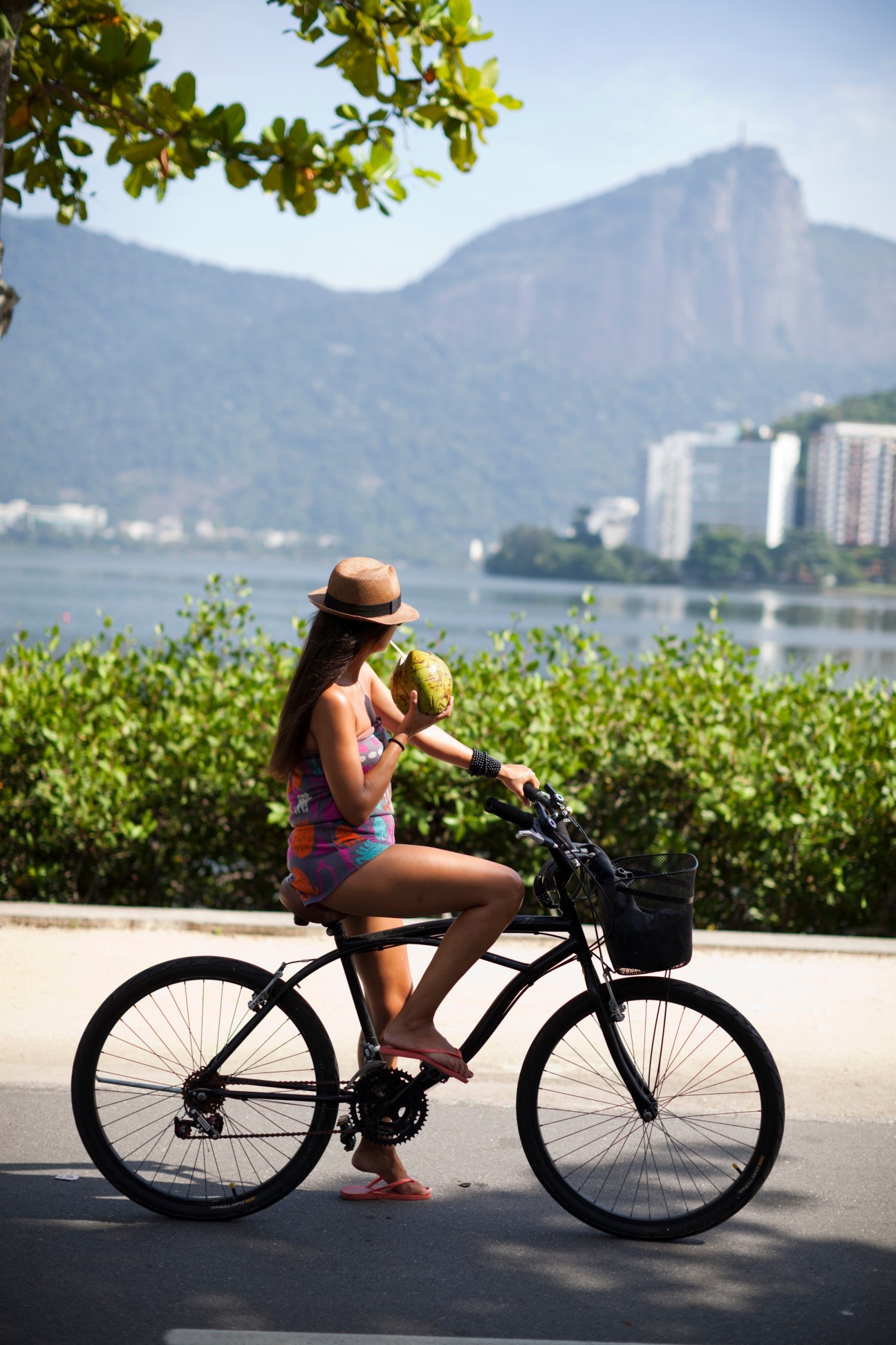 Woman on bicycle holding a coconut near Lagoa Rodrigo de Freitas in Rio de Janeiro with mountains in background.