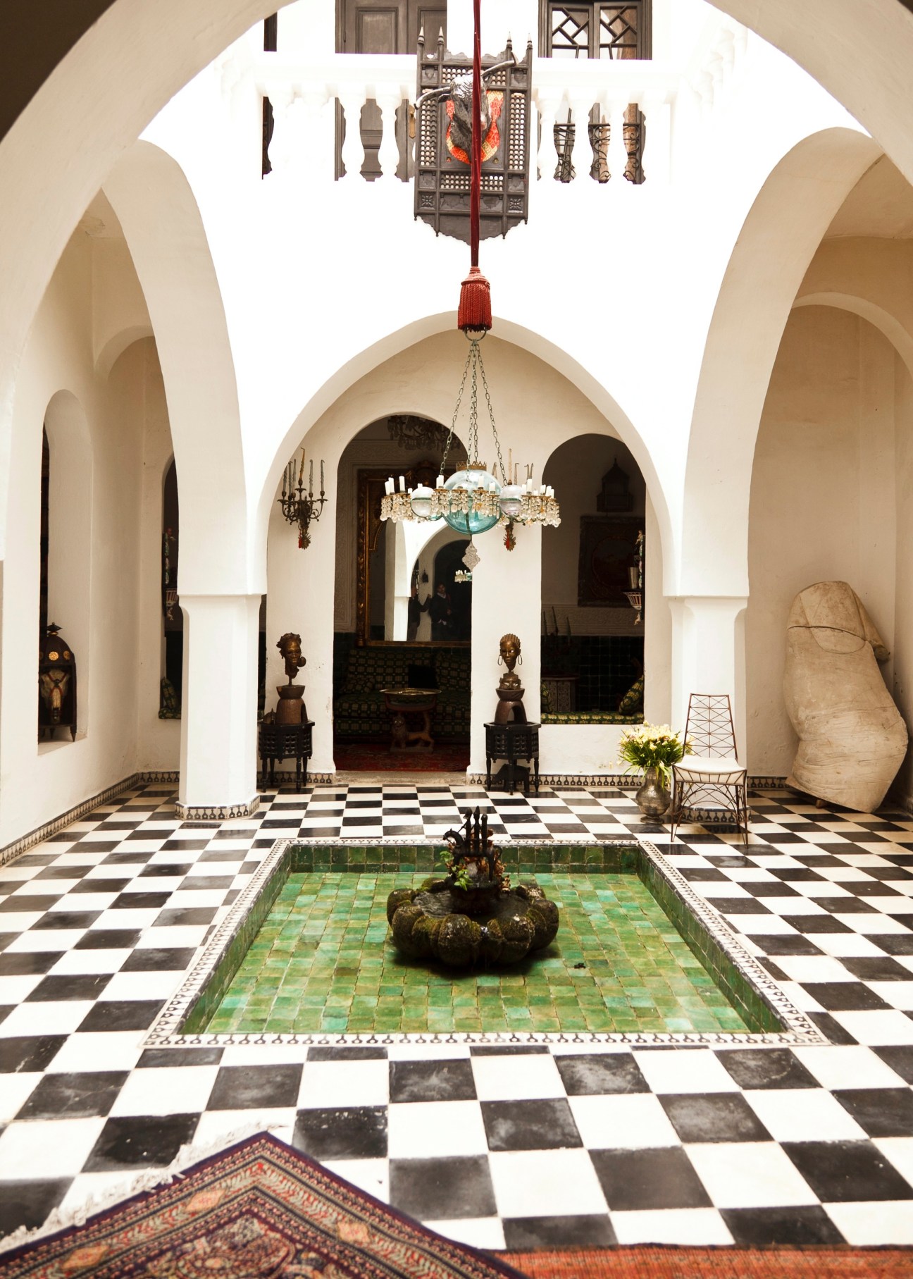 Grand courtyard of a traditional Moroccan riad with arched white walls, black and white checkered floor, central fountain, ornate chandelier, and decorative sculptures.