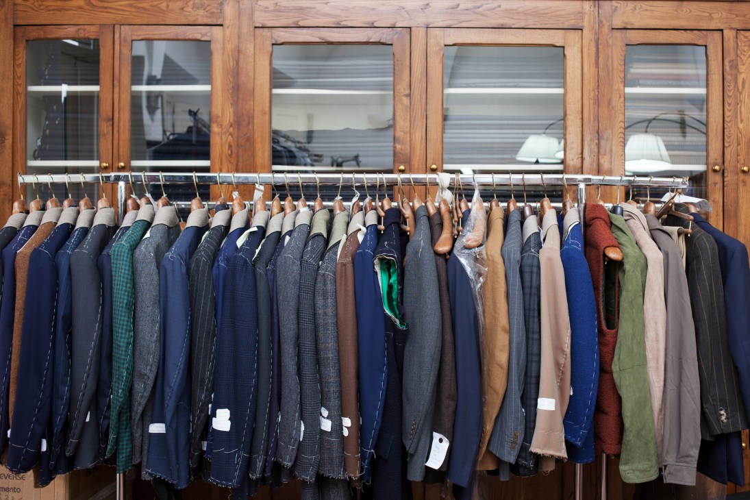 Row of completed tailored jackets in various colors and fabrics hanging on a metal rod in front of wooden cabinets.