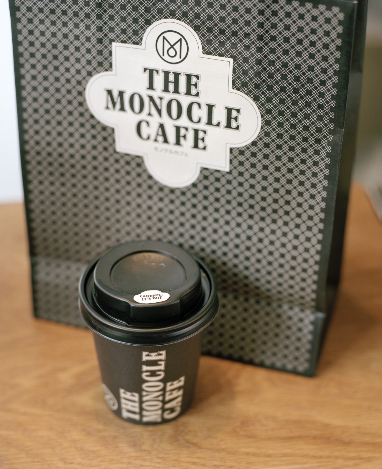 Monocle Café branded paper cup and shopping bag with logo on wooden surface.