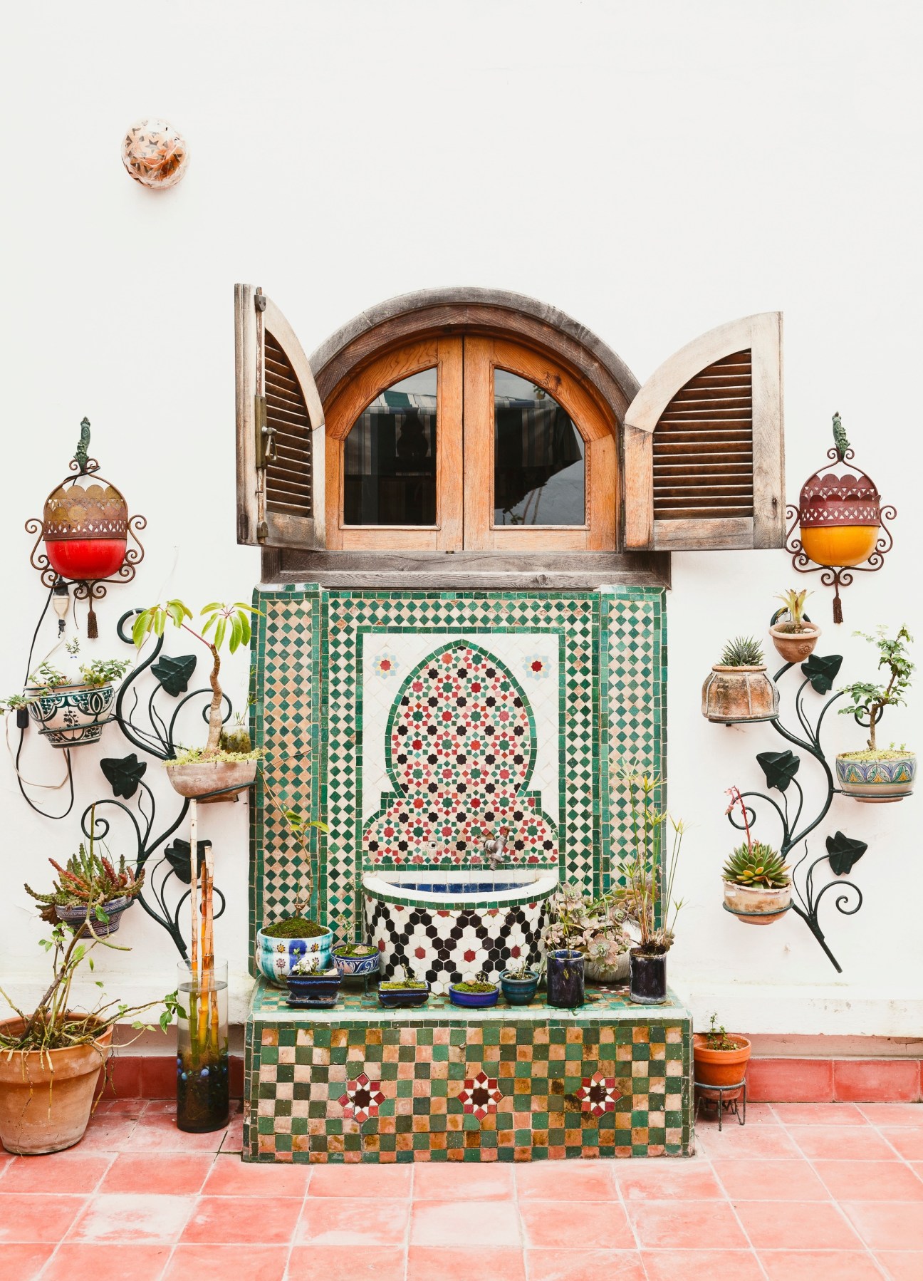 Ornate Moroccan courtyard fountain with zellige tilework, arched wooden window shutters, and potted plants on terracotta tiles.