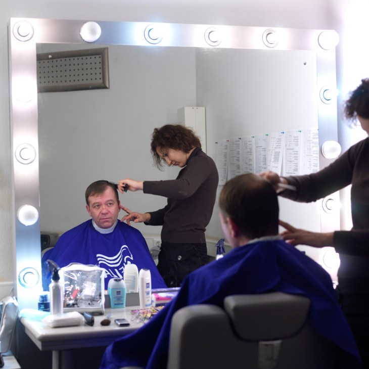 Peter Lavelle sits in a makeup chair while a makeup artist styles his hair in a brightly lit mirror at Russia Today studios.