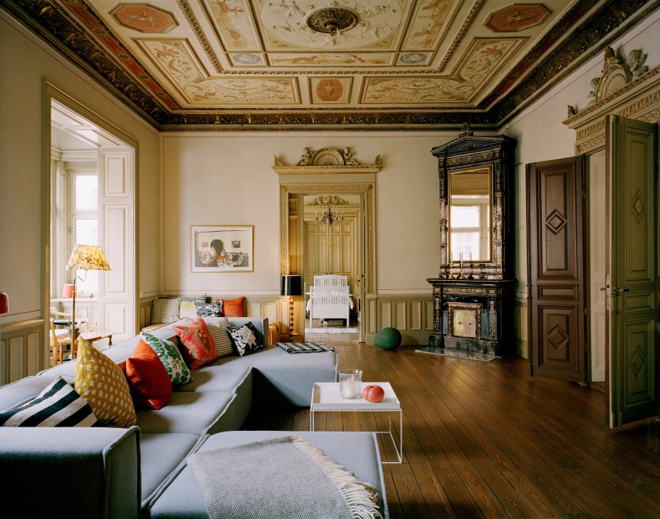 Ornate living room with painted ceiling, hardwood floors, green doors, fireplace and modern grey sofa with colorful cushions.