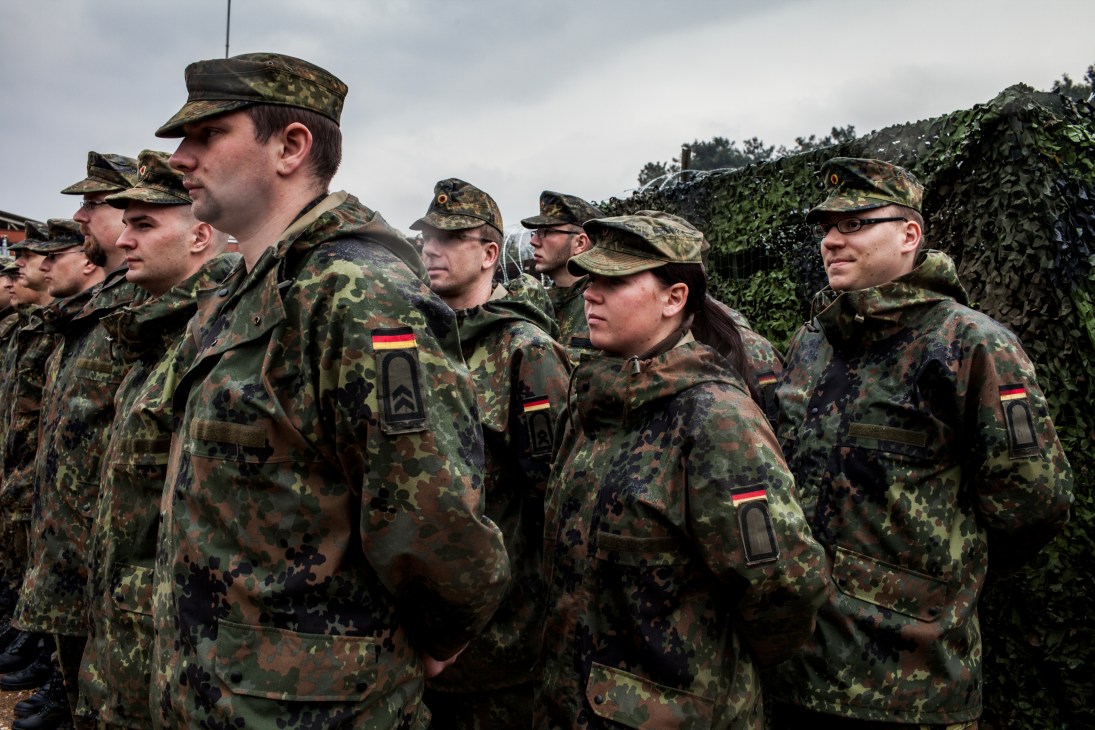 German military personnel in camouflage uniforms stand in formation outdoors.