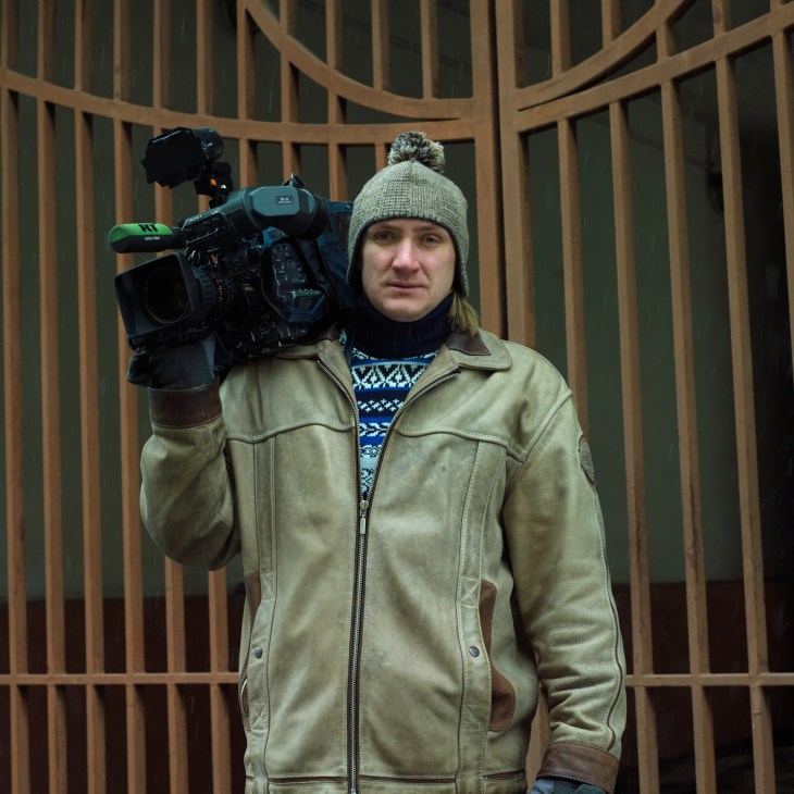 Cameraman Vladimir Batalin holds a professional video camera on his shoulder while wearing a beige jacket and gray beanie.