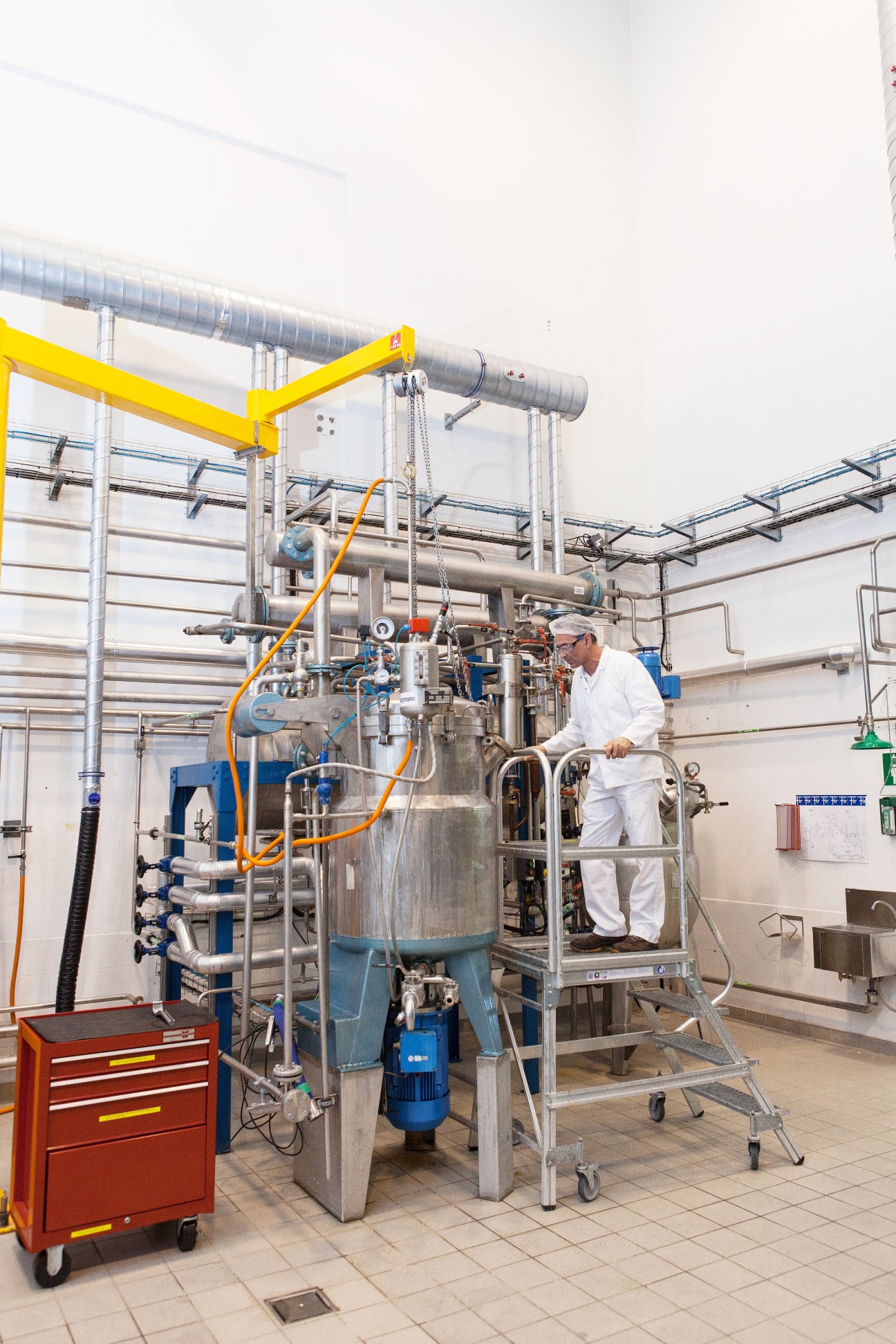 Worker in white protective clothing operates a large industrial fractionating column with yellow overhead crane at Firmenich Grasse facility.