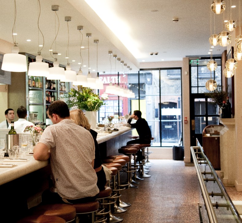 Interior of Bocca di Lupo restaurant showing a counter with diners seated on brown leather stools and pendant lighting overhead.