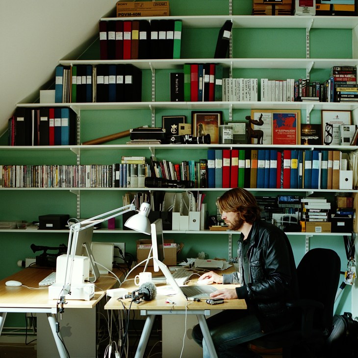Ruben Östlund sits at a desk in front of green shelving filled with books, films, and design materials in a creative studio workspace.