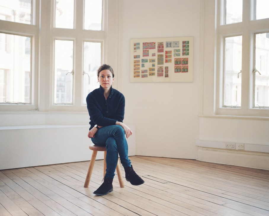 Hannah Barry sits on a wooden stool in a bright gallery space with large windows and framed artwork on the wall behind her.