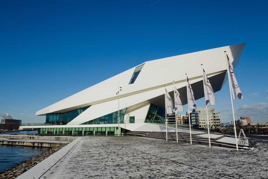 Modern white curved building with glass windows on Amsterdam waterfront with flags and blue sky.