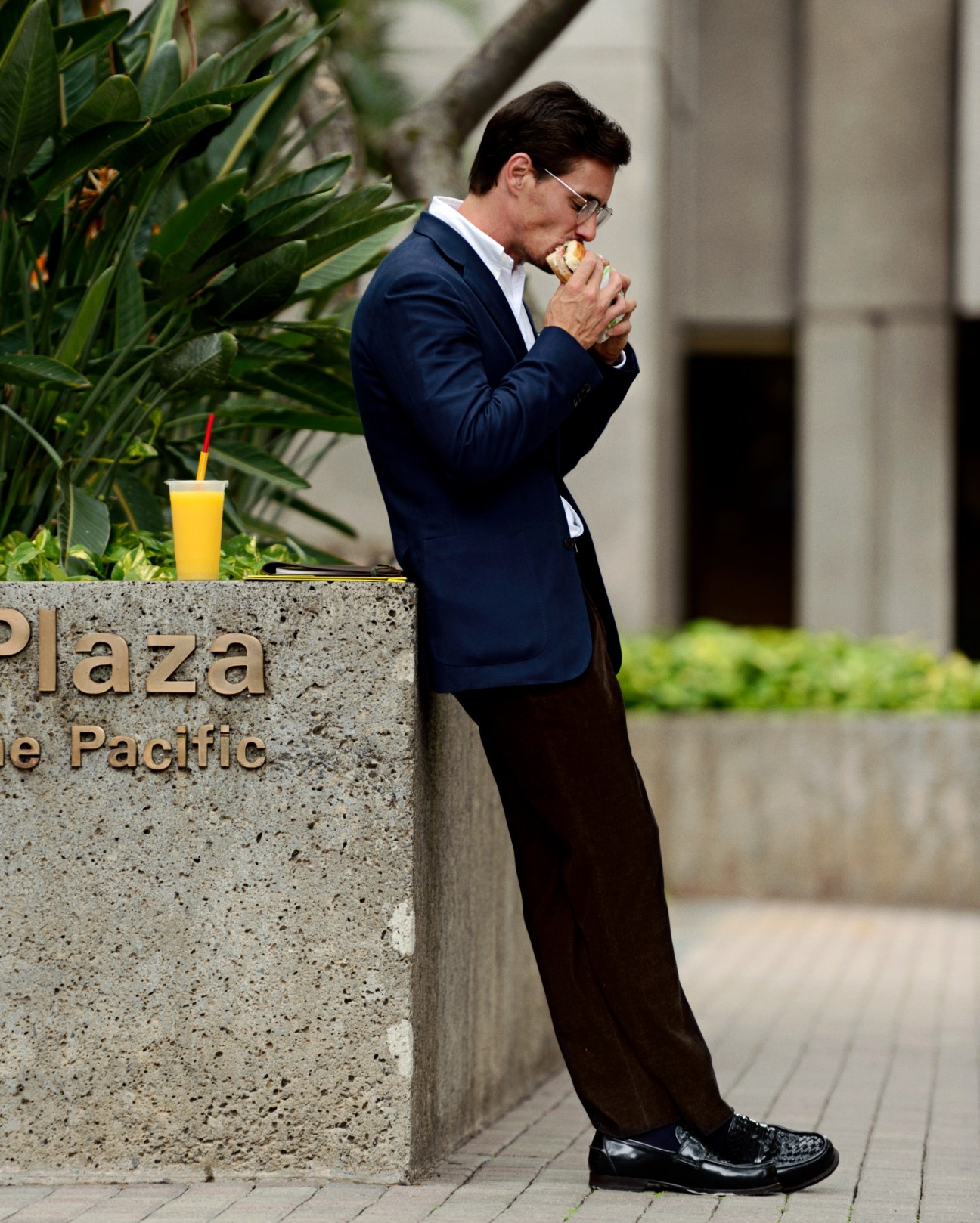 Man in navy coat and dark trousers eating a sandwich outdoors near tropical plants.