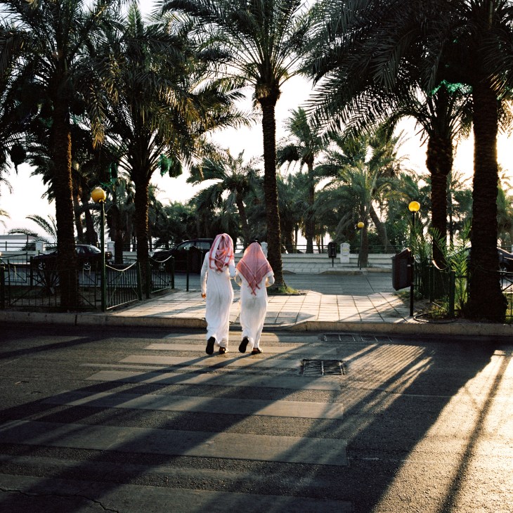 Two locals in white traditional dress walk along a palm-lined waterfront promenade in Aqaba.