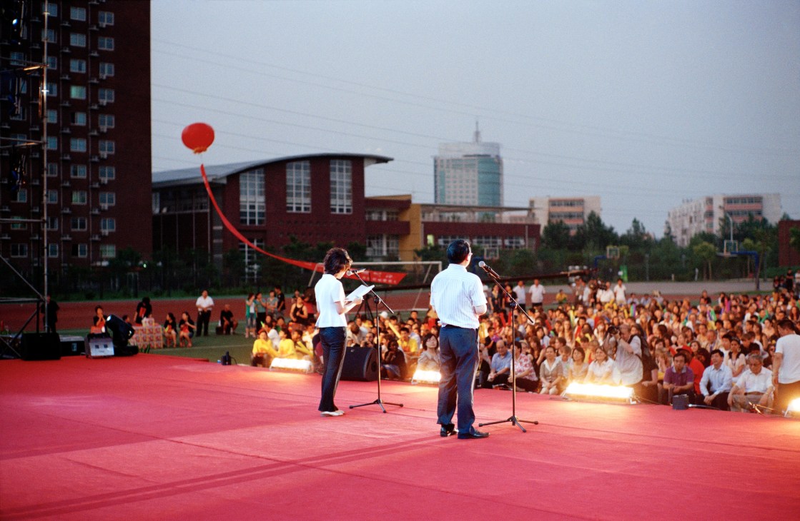 Two speakers address a large crowd from a red stage at the Chinese Bridge summer camp opening ceremony in Beijing.