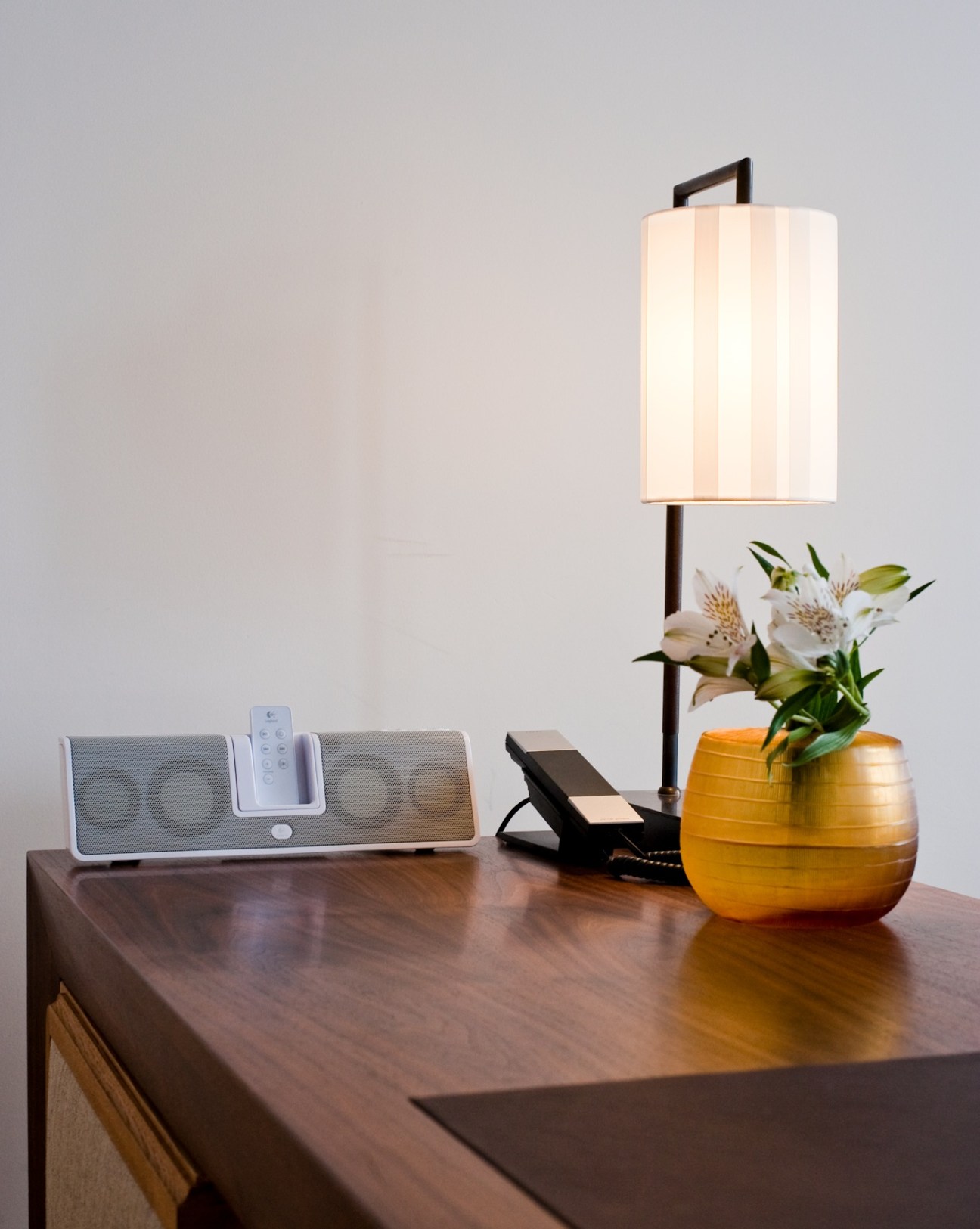 Modern desk setup with speaker system, lamp, and gold vase with white flowers on wooden table.