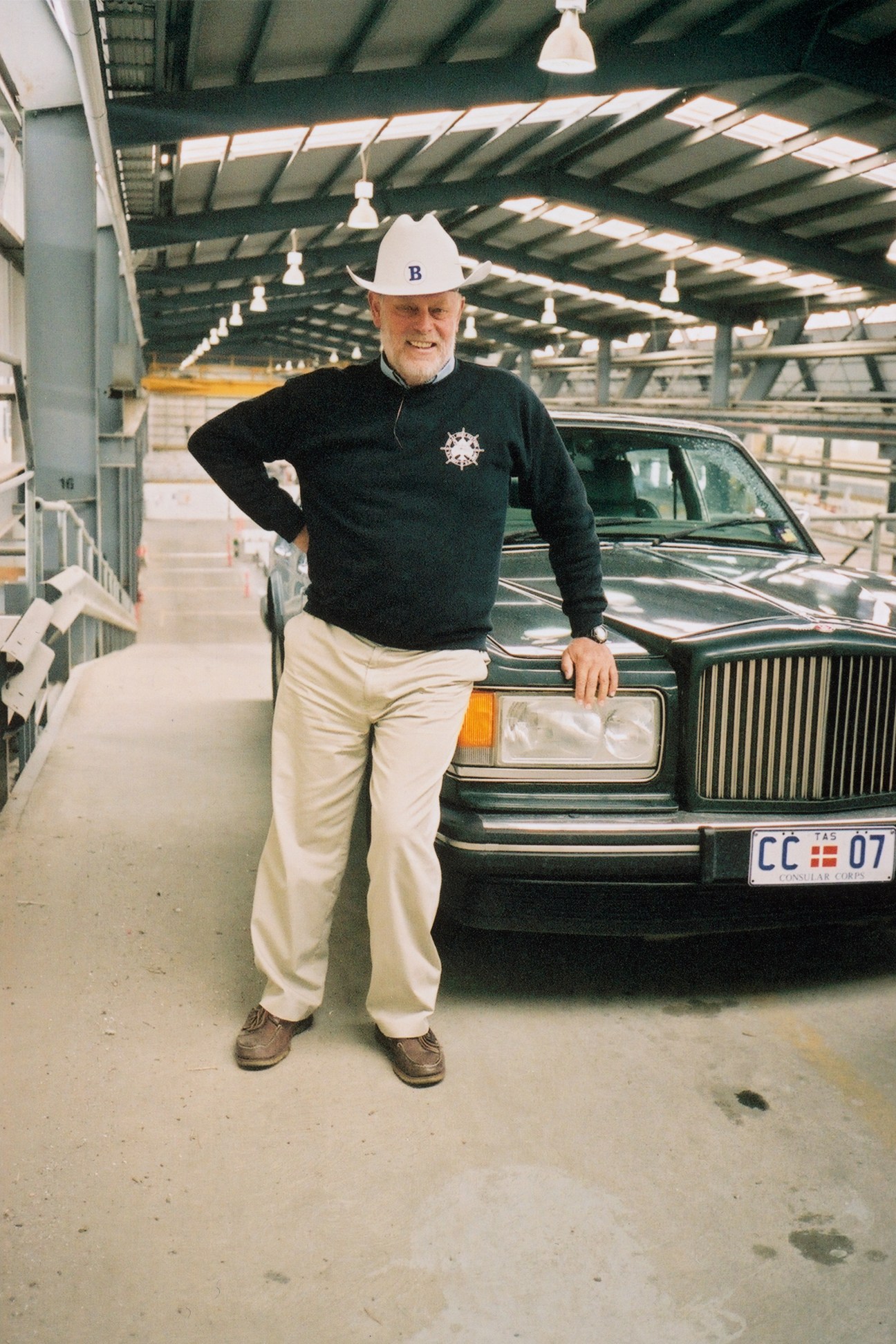 Robert Clifford, owner of INCAT, stands in a shipyard wearing a white hard hat and black sweater with a ship's wheel logo, next to a green car.