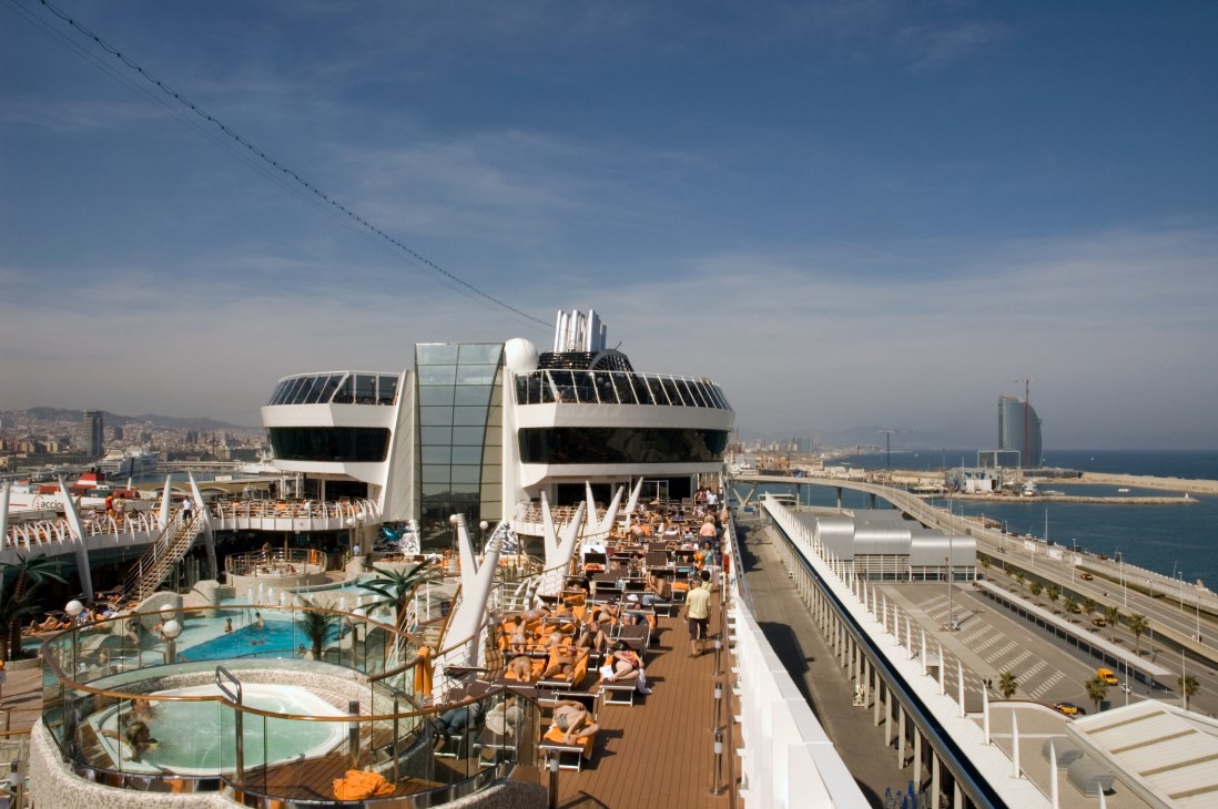 Passengers relax on the sun deck of MSC Fantasia cruise ship with Barcelona port visible in background.