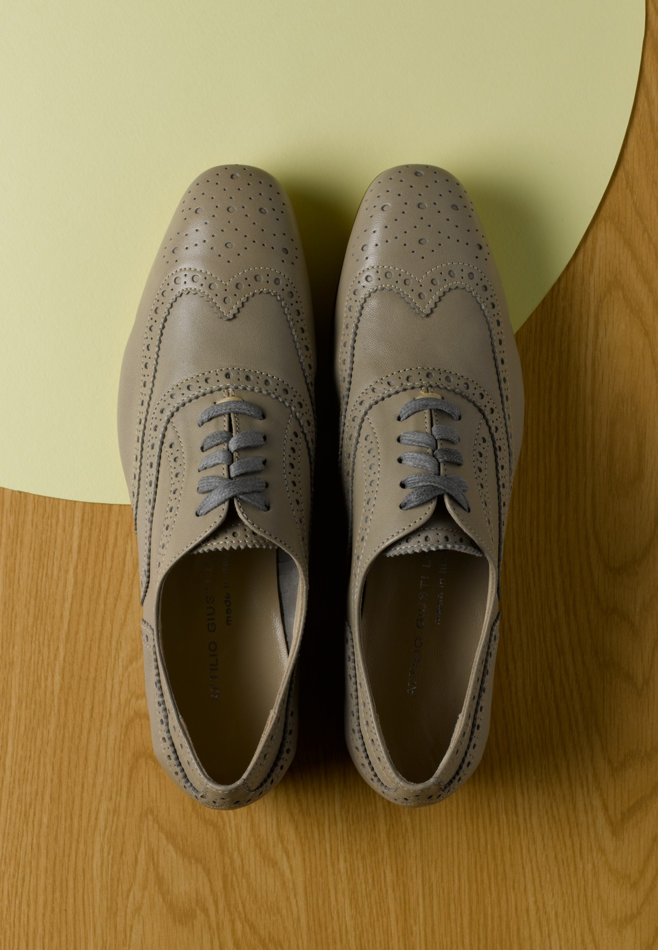Pair of beige lambskin nappa brogues with perforated detailing and dark laces, displayed on a wooden floor.