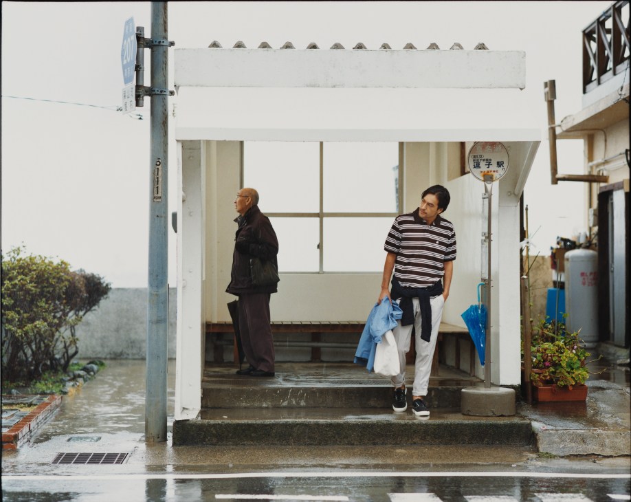 Two men in a bus shelter in rainy Hayama, Japan; one wearing a black coat, the other in striped shirt and white trousers holding a blue jacket.
