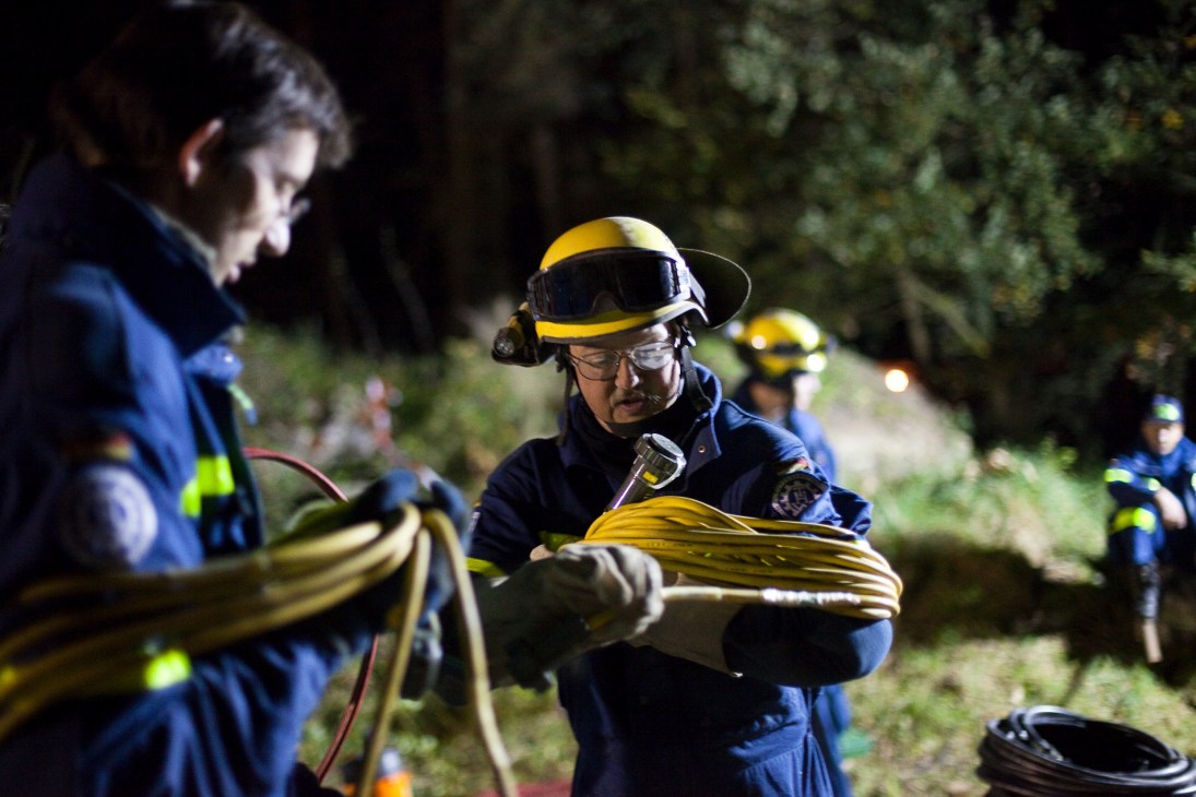THW volunteer in blue uniform and yellow helmet handles coiled cable during nighttime rescue training operation.