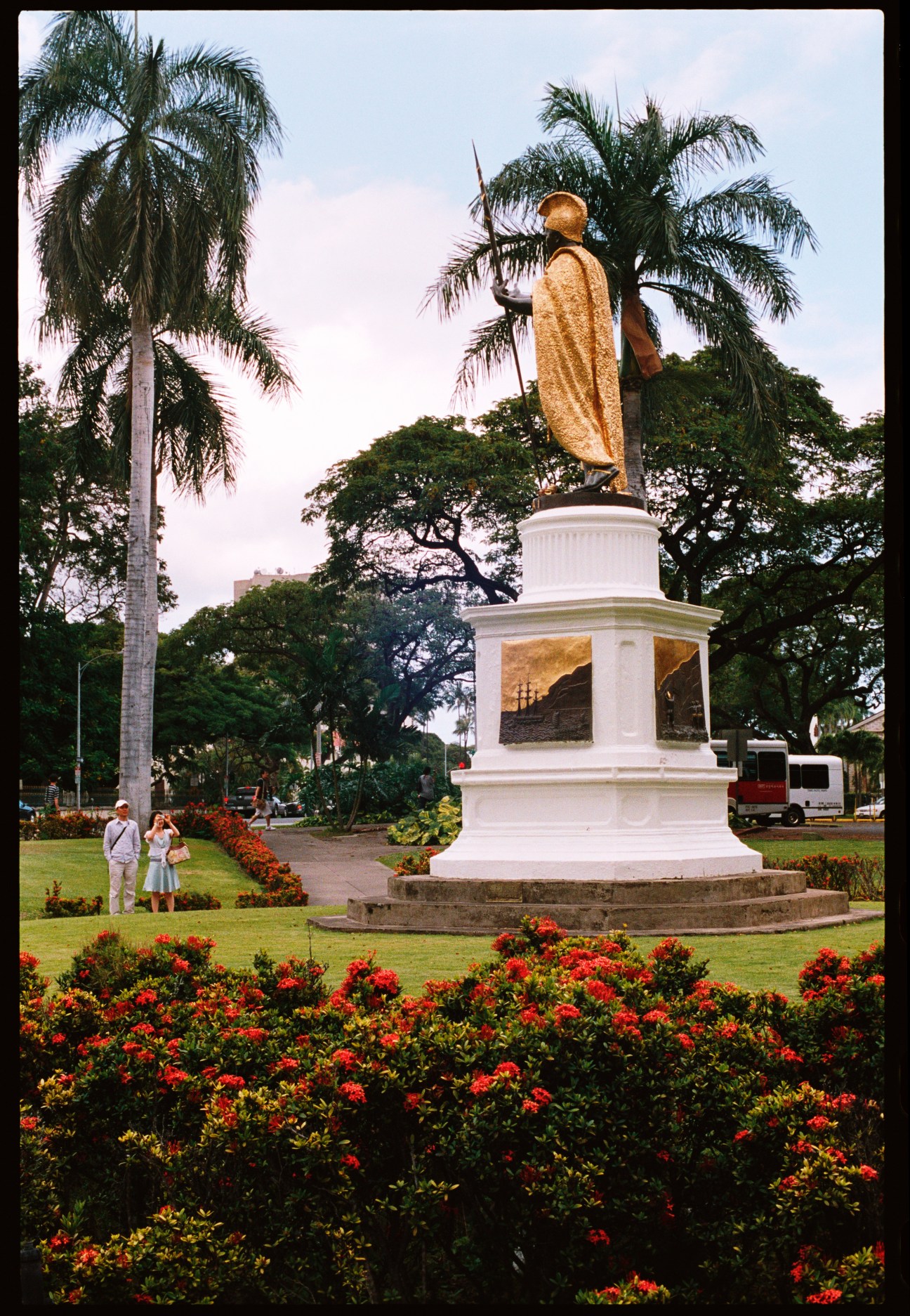 A statue of King Kamehameha in a white pedestal monument surrounded by red flowers and palm trees.