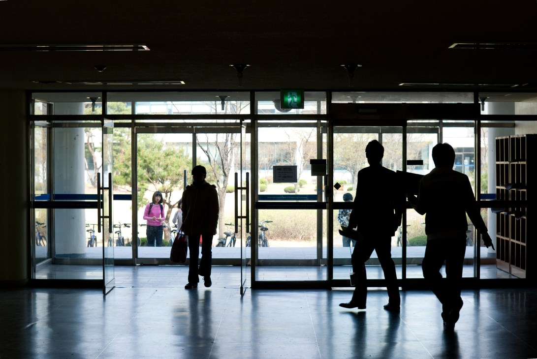 Silhouetted figures walking through glass doors of a modern KAIST building entrance.