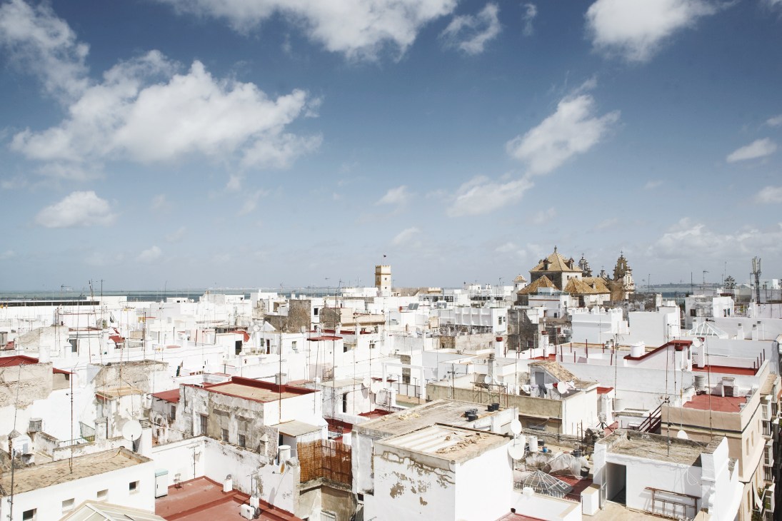 Aerial view of Cádiz's historic Casco Antiguo district with white buildings, red-tiled roofs, and cathedral domes under blue sky.