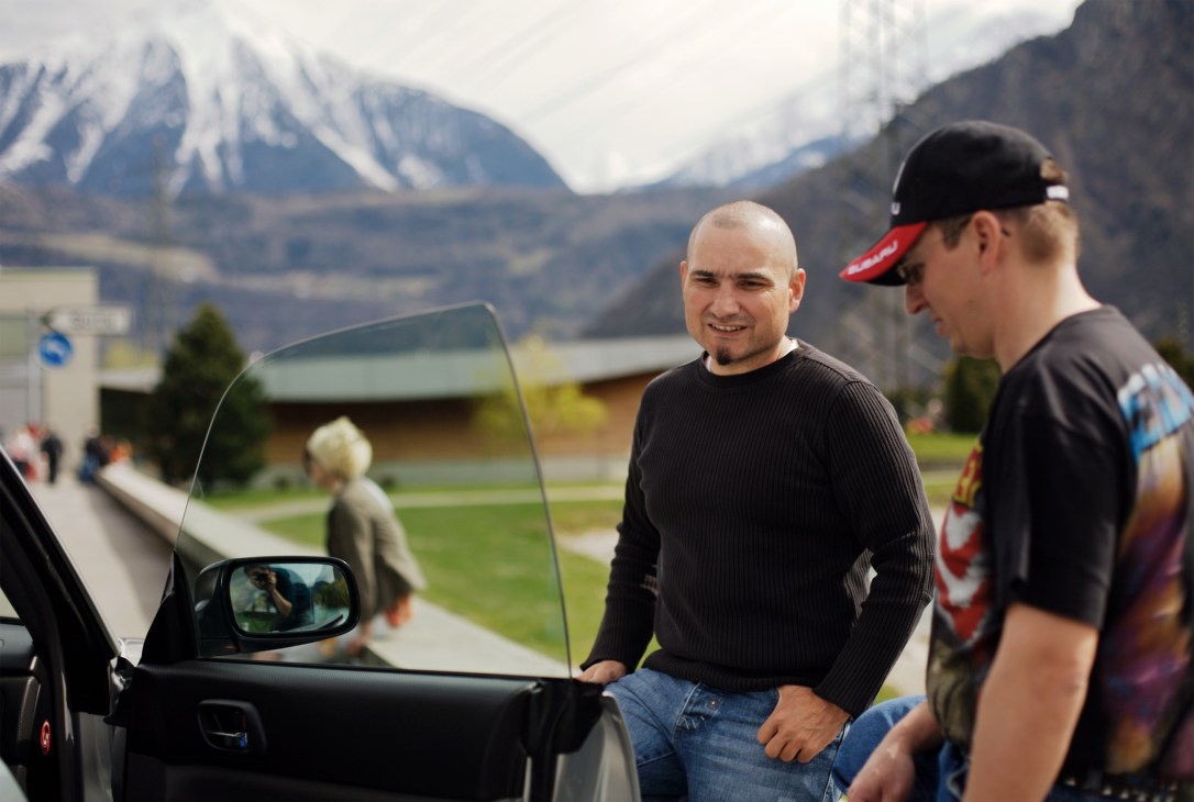 Two men chat beside a Subaru in a Swiss Alpine valley with snow-capped mountains in the background.