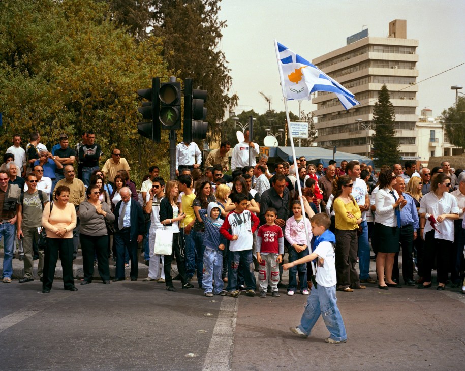 A crowd celebrates Greek Independence Day in South Cyprus, with a Cypriot flag visible among gathered citizens and children.