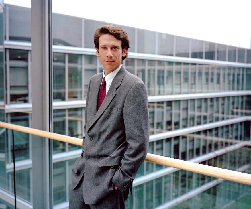 Konstantin Neven DuMont in grey suit and red tie standing on modern building balcony.