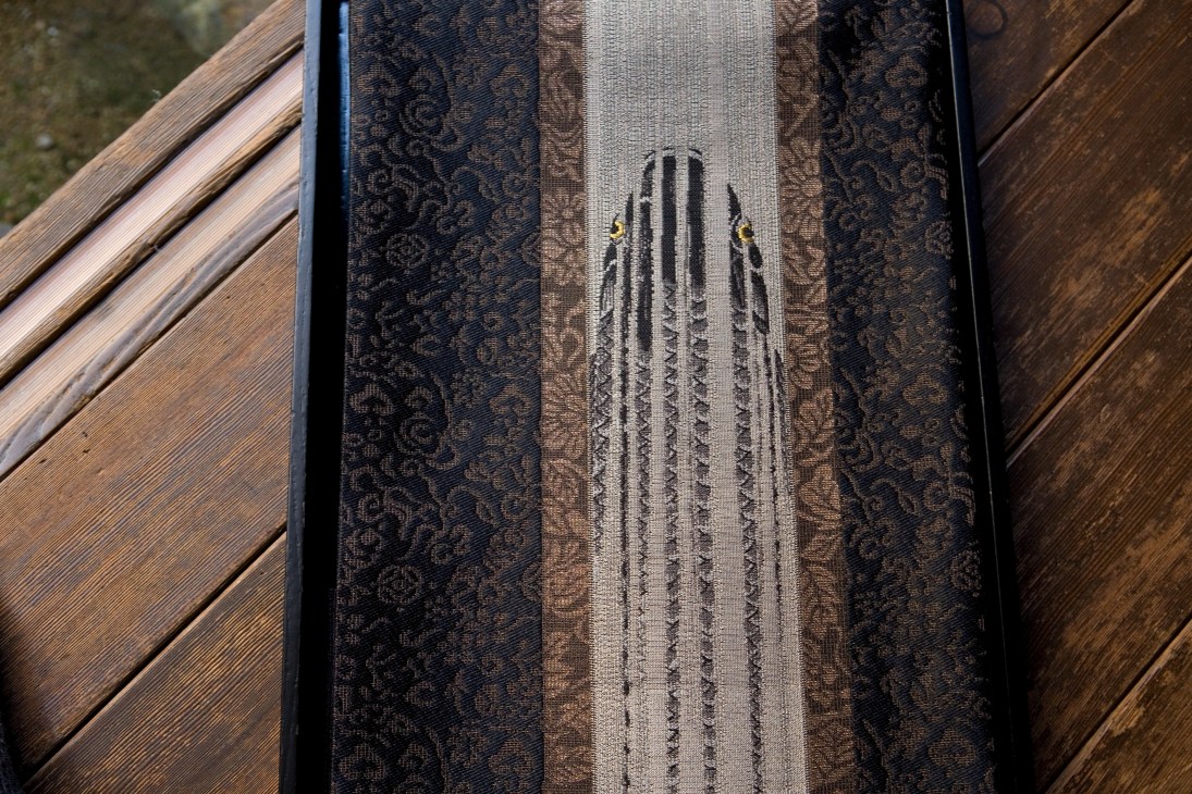 Close-up of woven textile showing a carp design in gray and dark patterned fabric strips on wooden surface.