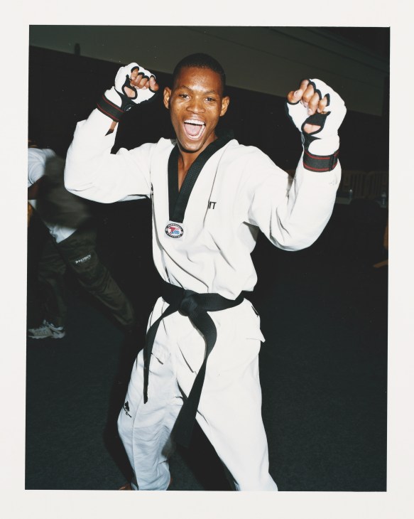 Male taekwondo athlete from Mozambique in white uniform with black belt, raising both fists in victory pose.