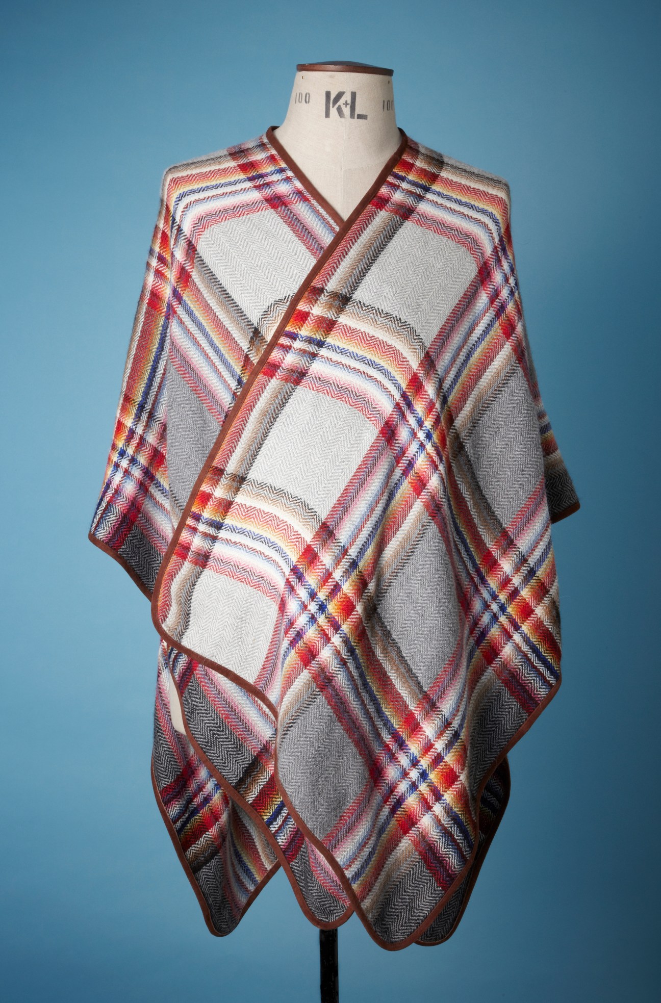 Inoue travel cape with multicolored plaid pattern in red, blue, and cream displayed on dress form against blue background.