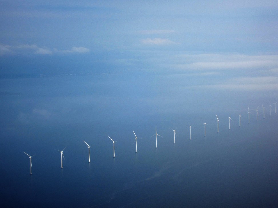Aerial view of offshore wind turbines in a line across blue water beneath a cloudy sky.