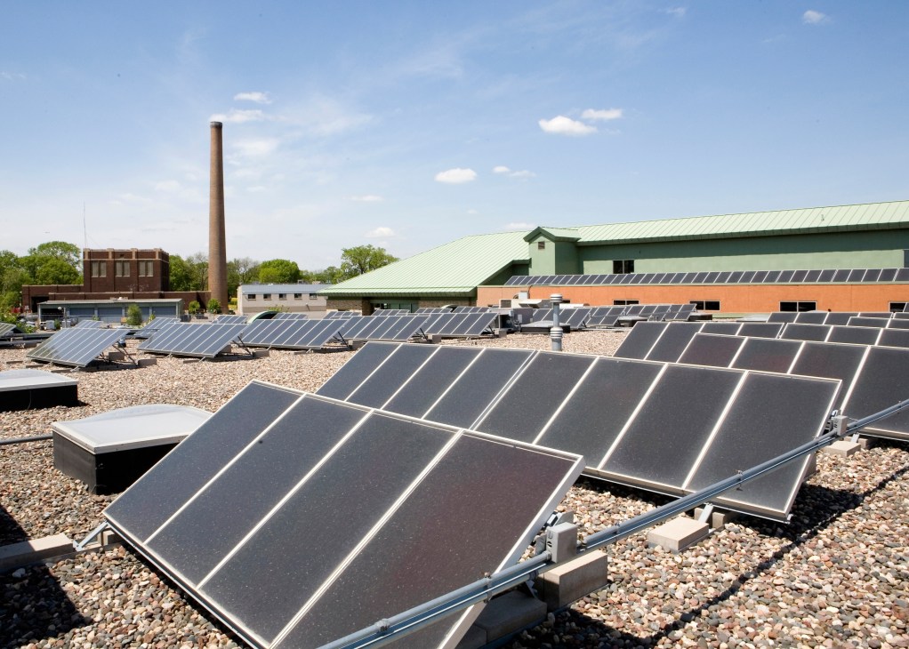 Solar panels on a gravel rooftop with industrial buildings and a smokestack in the background.