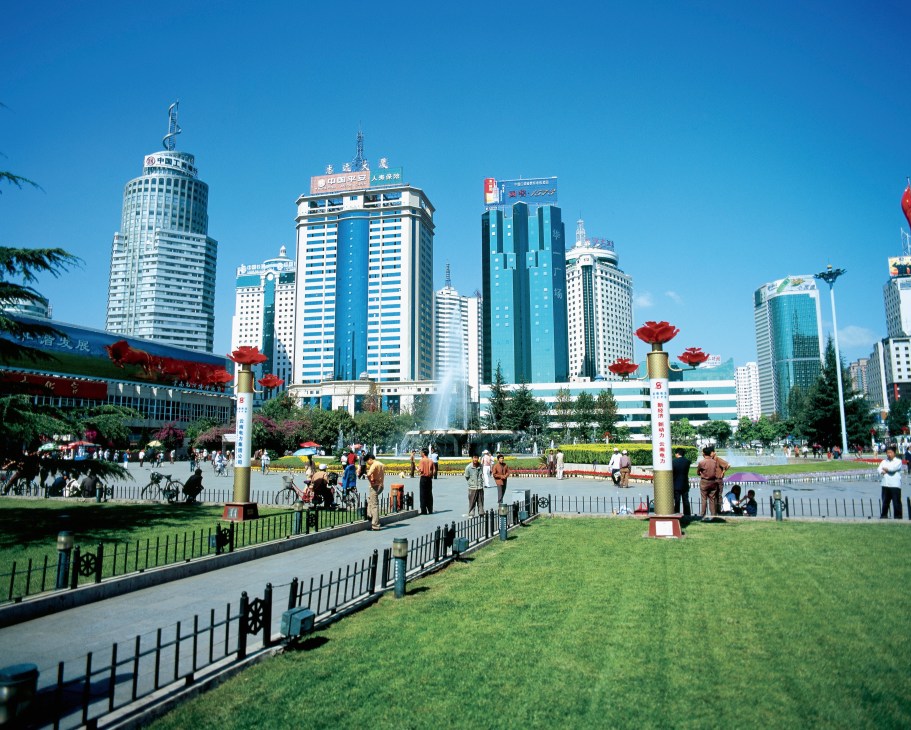 A public plaza in Kunming with modern skyscrapers, green lawn, red lantern decorations, and people walking under a clear blue sky.