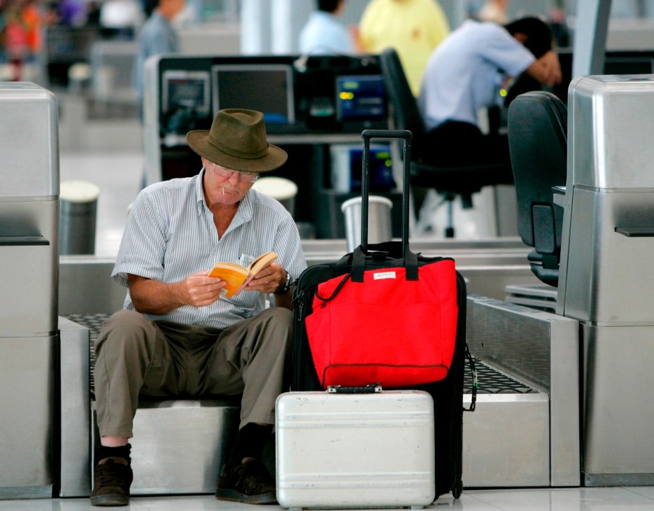 Man in hat sitting at airport gate with luggage and documents.