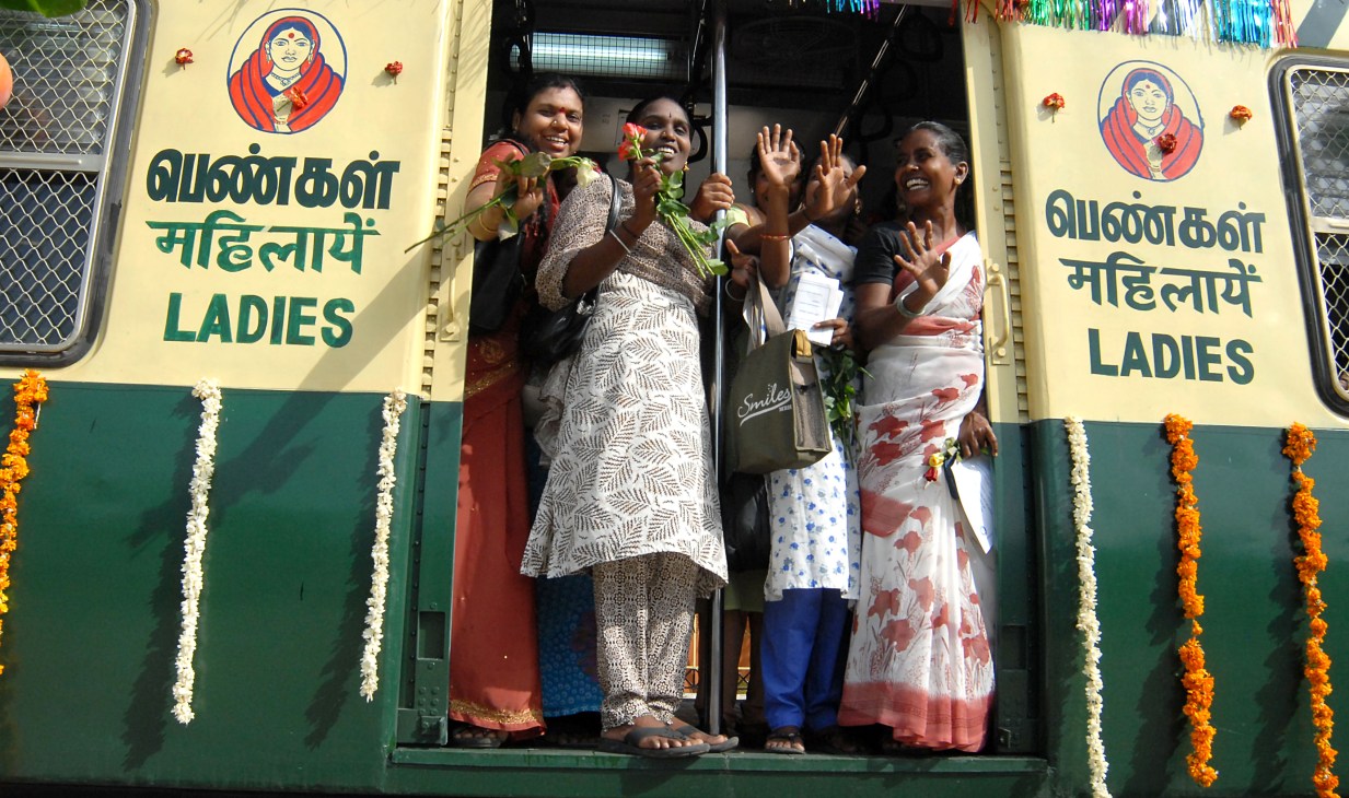 Women waving from the doorway of a yellow and green train car marked "LADIES" in multiple languages.