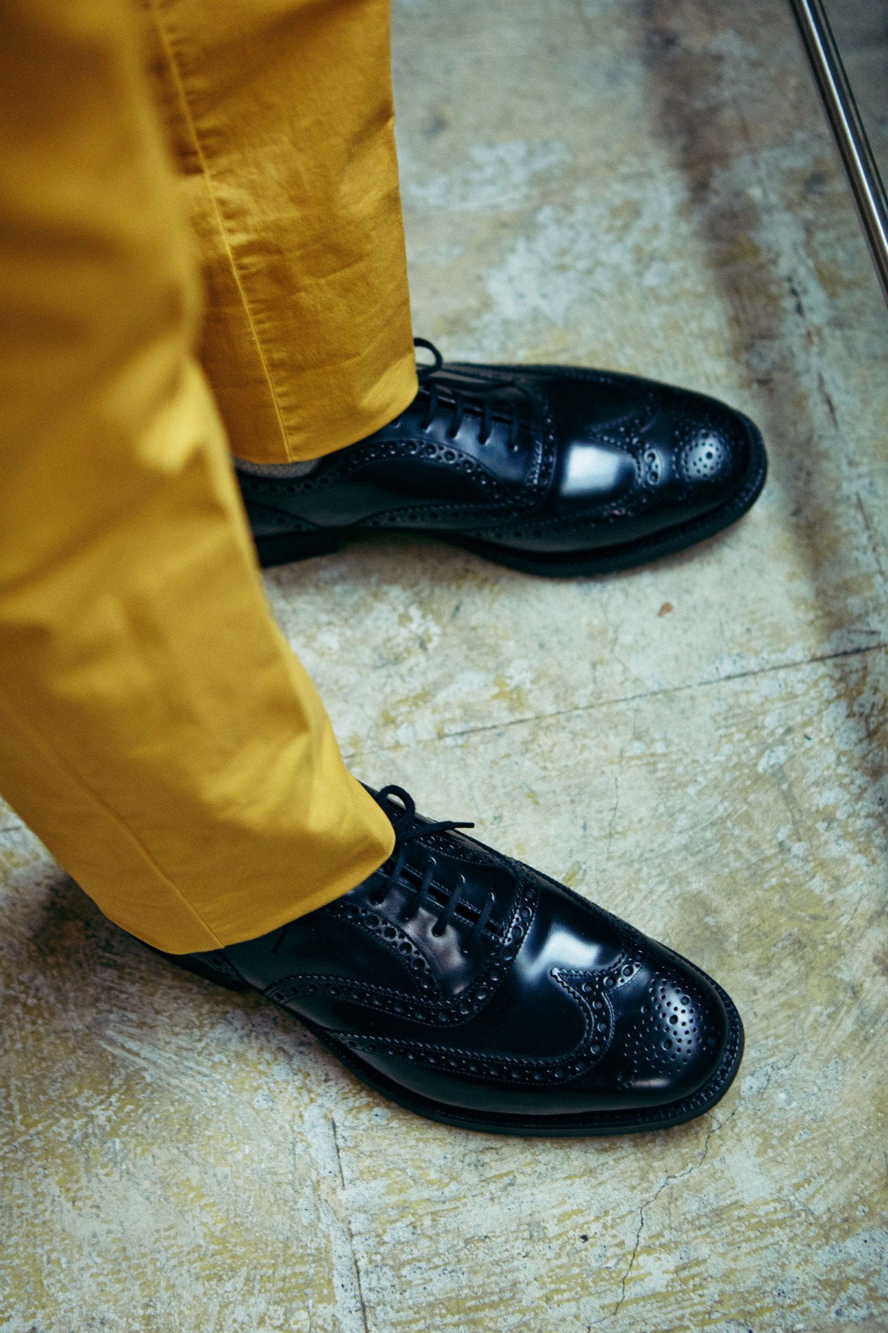 Person wearing mustard-yellow trousers by Germano for Edifice and black brogue shoes by Church's on a concrete floor.