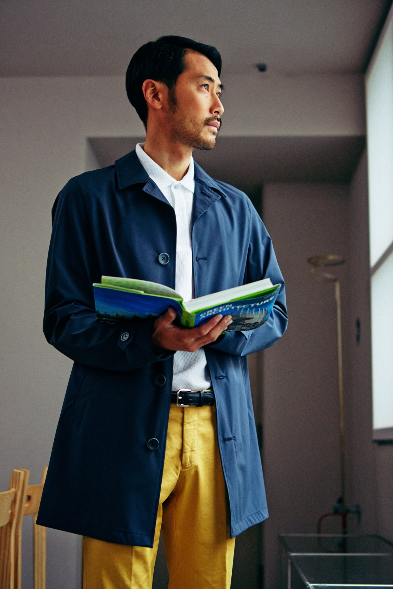 Man wearing navy Herno coat, white shirt, yellow Germano trousers, and black belt holding an open book.