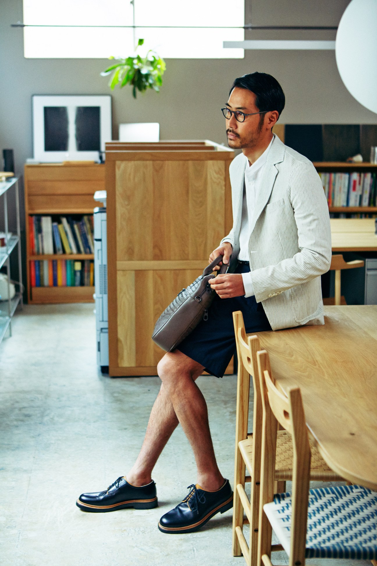 Man in light jacket, shorts and Valentino Garavani shoes holding Bottega Veneta bag in modern office setting.