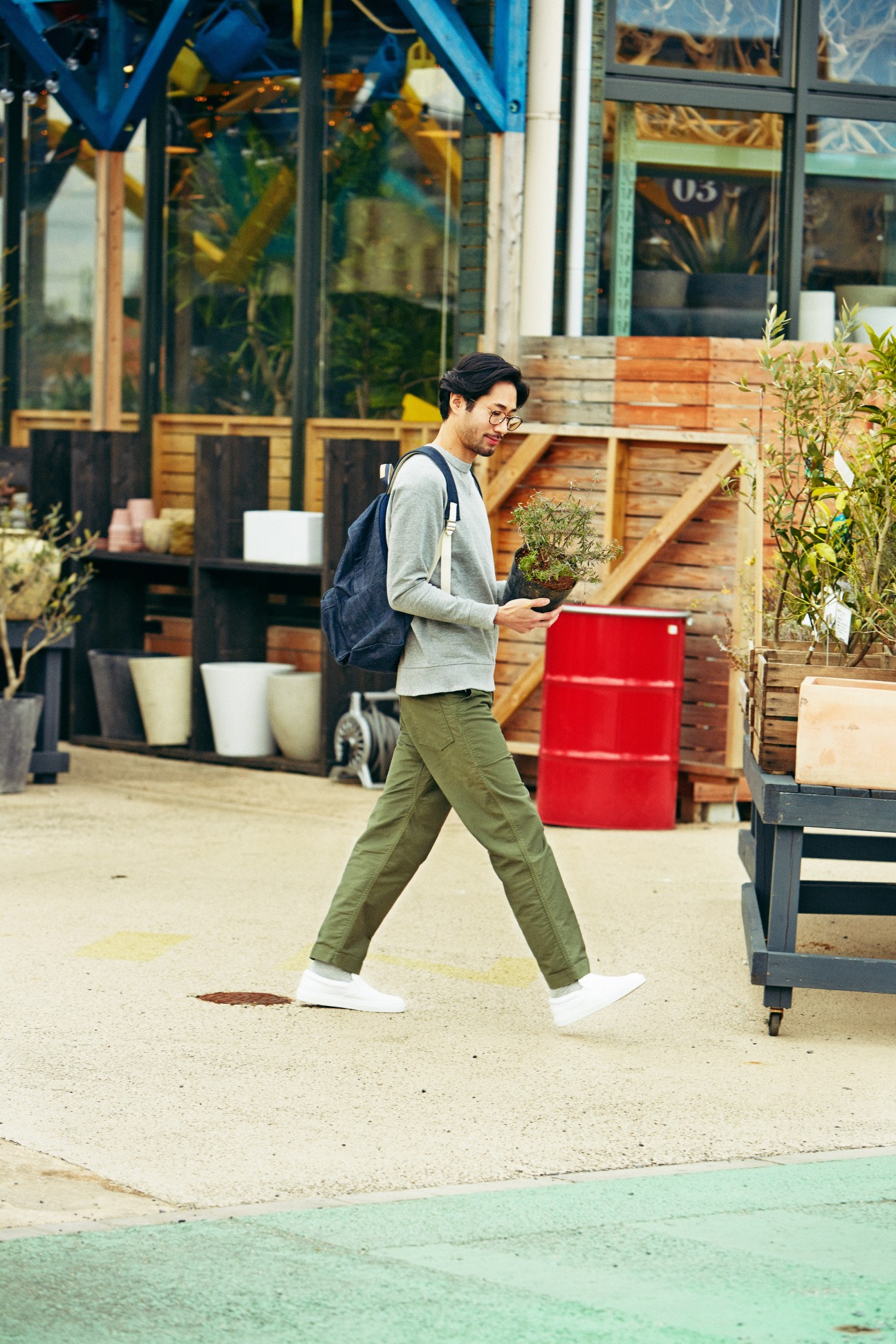 Man wearing grey sweatshirt, olive trousers, white socks, slip-on shoes and backpack walking outdoors.