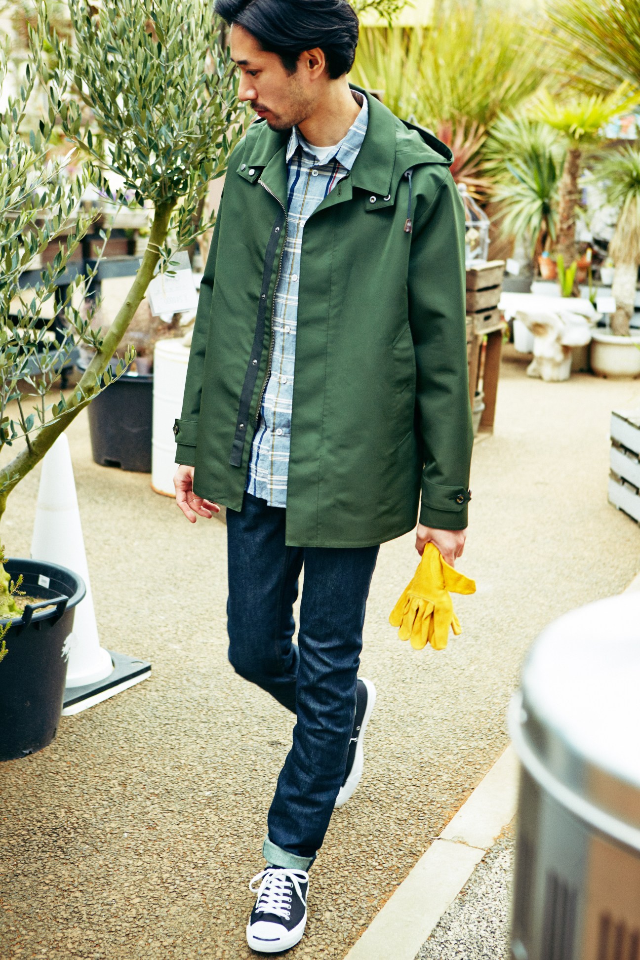 Man in green Sealup coat, Barena shirt, Louis Vuitton jeans and Converse trainers holding yellow gloves in a plant nursery.