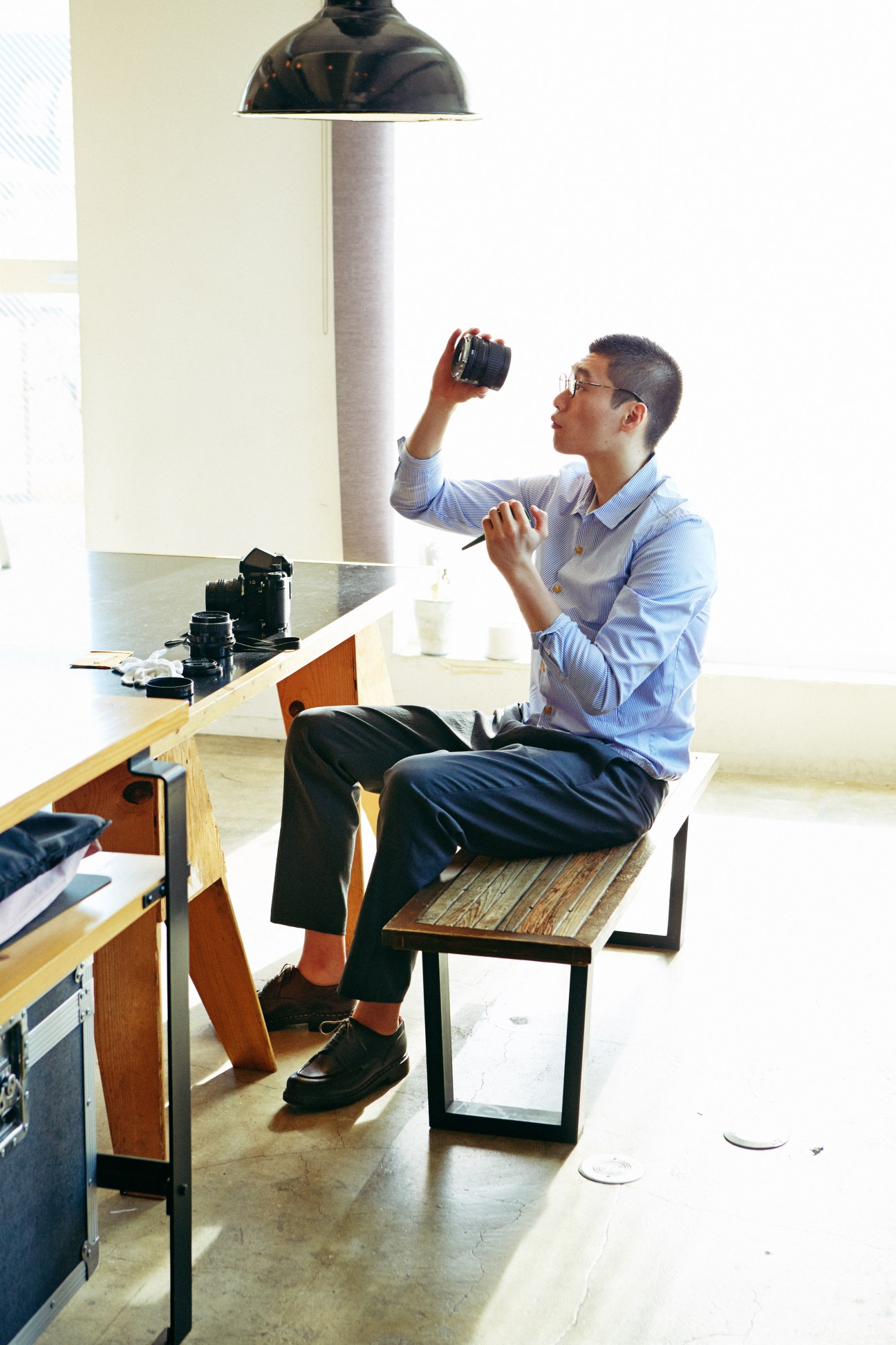Man in blue shirt and dark trousers sitting at desk, examining camera equipment.