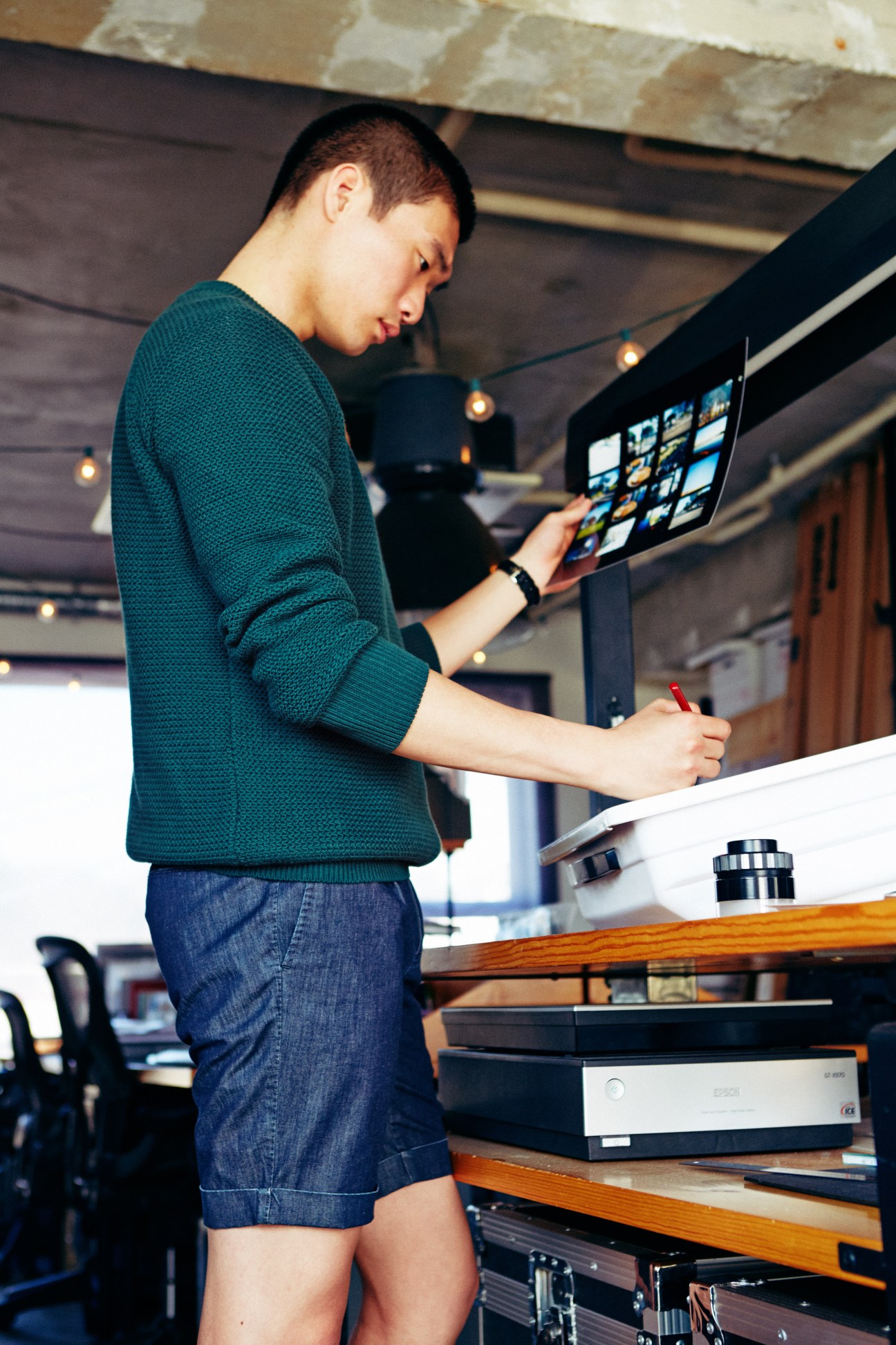 Man in teal knit jumper and denim shorts working at desk with tablet and laptop
