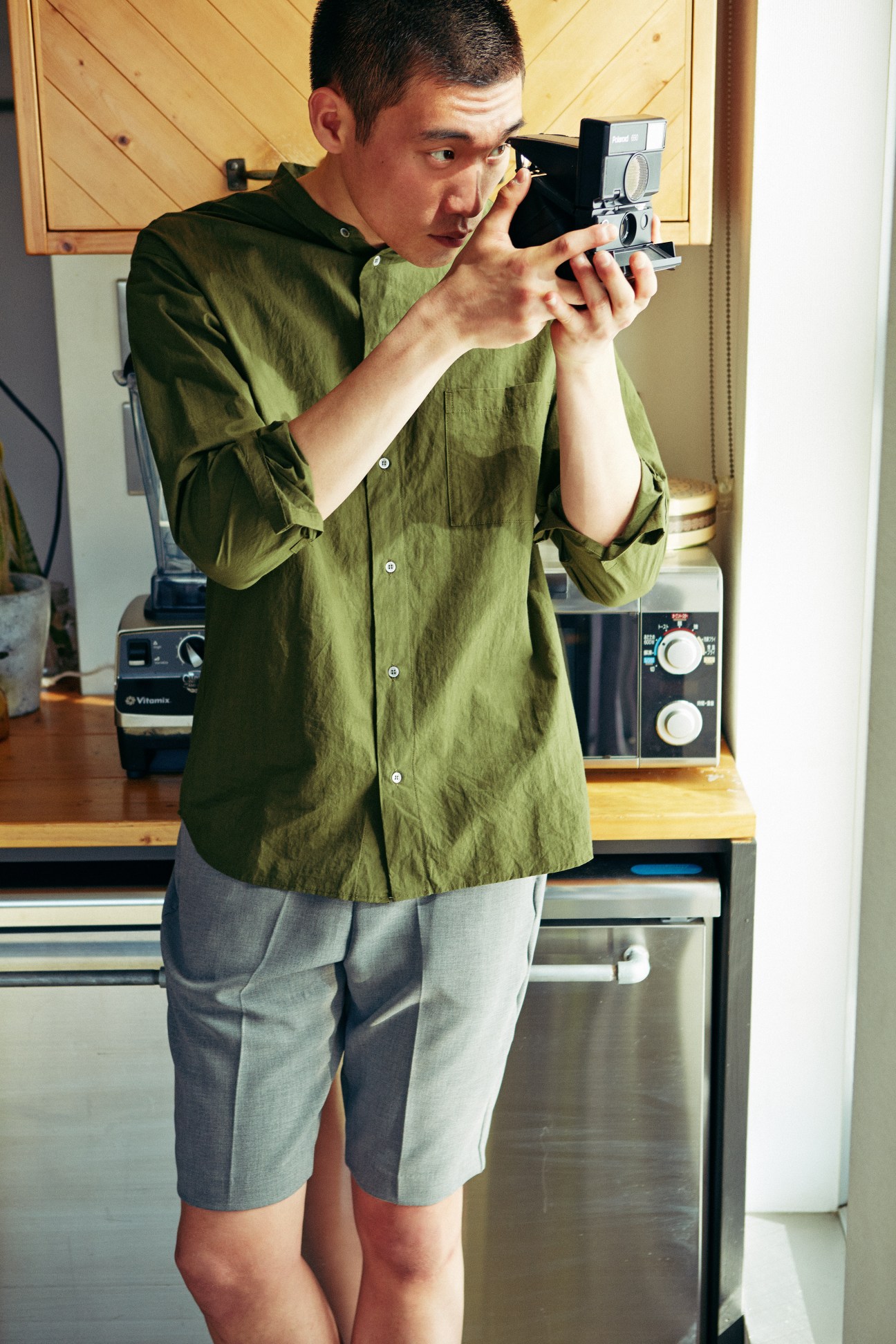 Man in olive shirt and grey shorts holding vintage film camera indoors.
