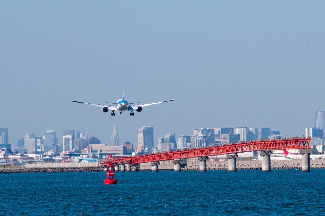 Commercial aircraft approaching over blue water with red bridge and city skyline in background.
