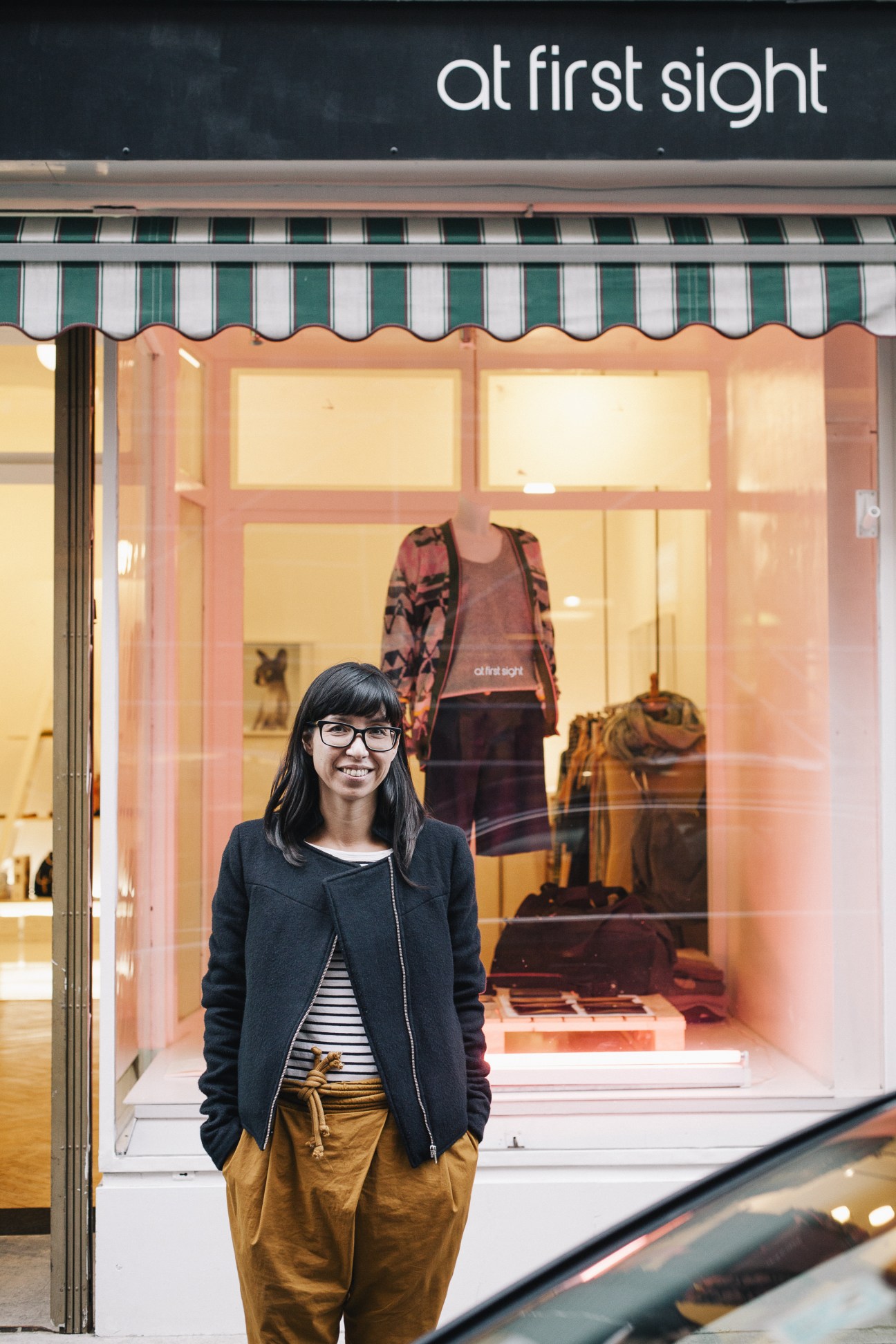 Woman with black hair and glasses standing in front of S/ght fashion store storefront.
