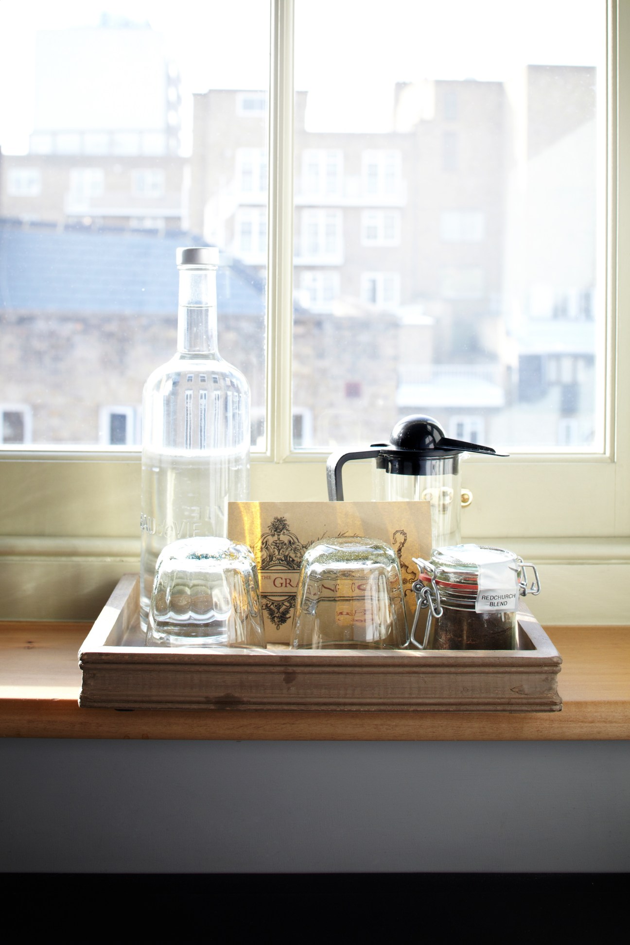 Hotel room minibar and beverage setup on wooden tray with city view through window.
