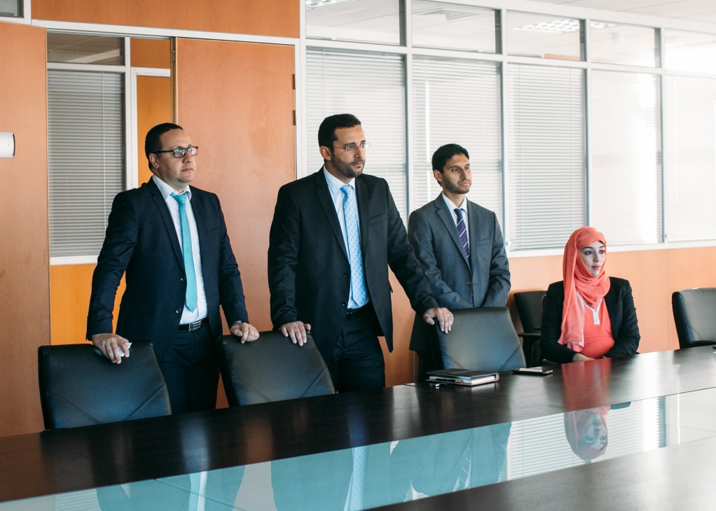 Four business professionals stand behind a conference table in a modern office with orange and white walls.