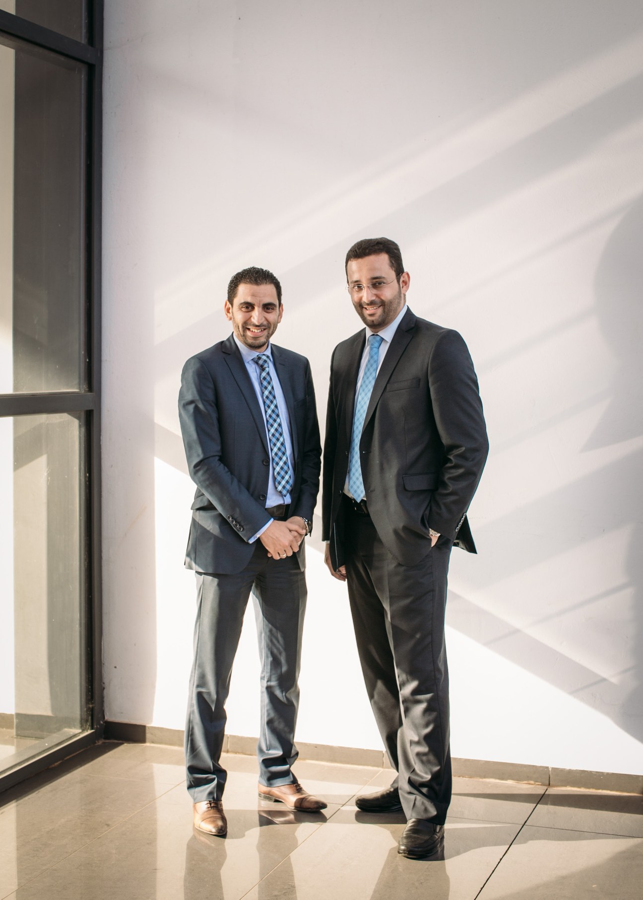 Two men in business suits shake hands in a minimalist white office space with dramatic window shadows.